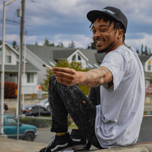 A man smiling next to a skateboard and passing a joint towards the camera