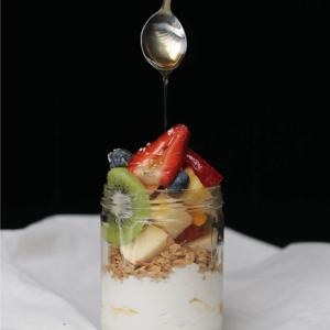 a spoon drizzling honey into a jar with yogurt, fruit, and granola