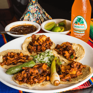 Tacos arranged in a circle next to limes, salsa, and a jarritos bottle