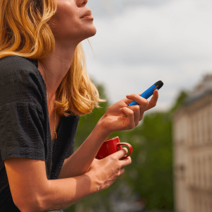 a woman drinking from a red mug and vaping on a balcony