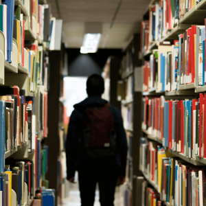 man with backpack walking through a library aisle lined with books
